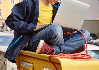 Junge sitzt mit Laptop und Kabelschnur auf einem Briefkasten